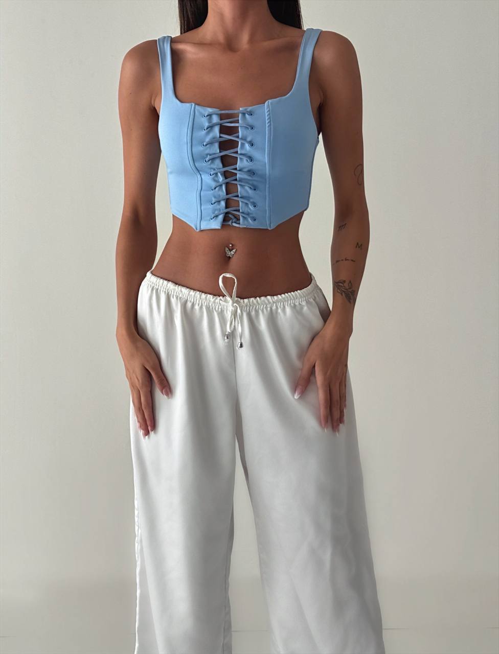 Woman wearing a blue lace-up crop top and white pants against a plain background