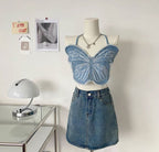 A mannequin wearing a blue denim crop top with a butterfly pattern, paired with a denim skirt. The setting includes a desk with a lamp and a photo frame in the background.