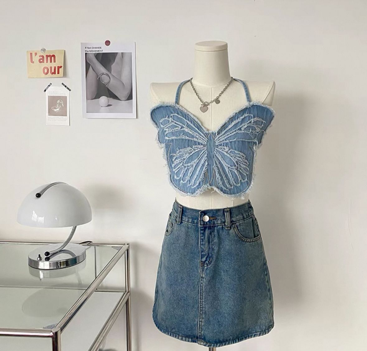 A mannequin wearing a blue denim crop top with a butterfly pattern, paired with a denim skirt. The setting includes a desk with a lamp and a photo frame in the background.