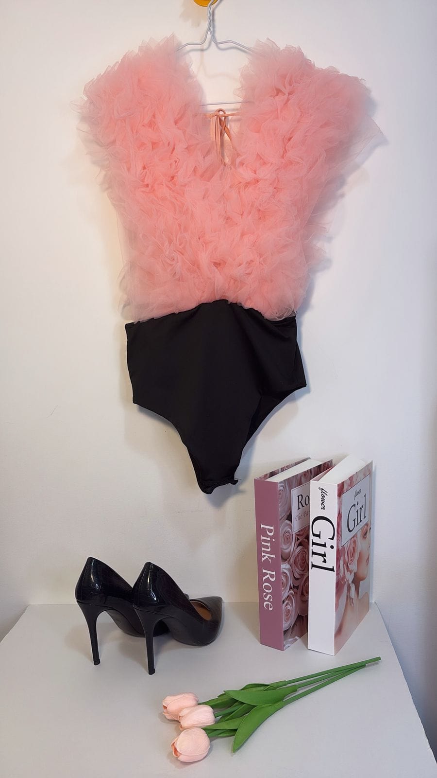 Pink bodysuit on a hanger with books and high heels on a white surface.