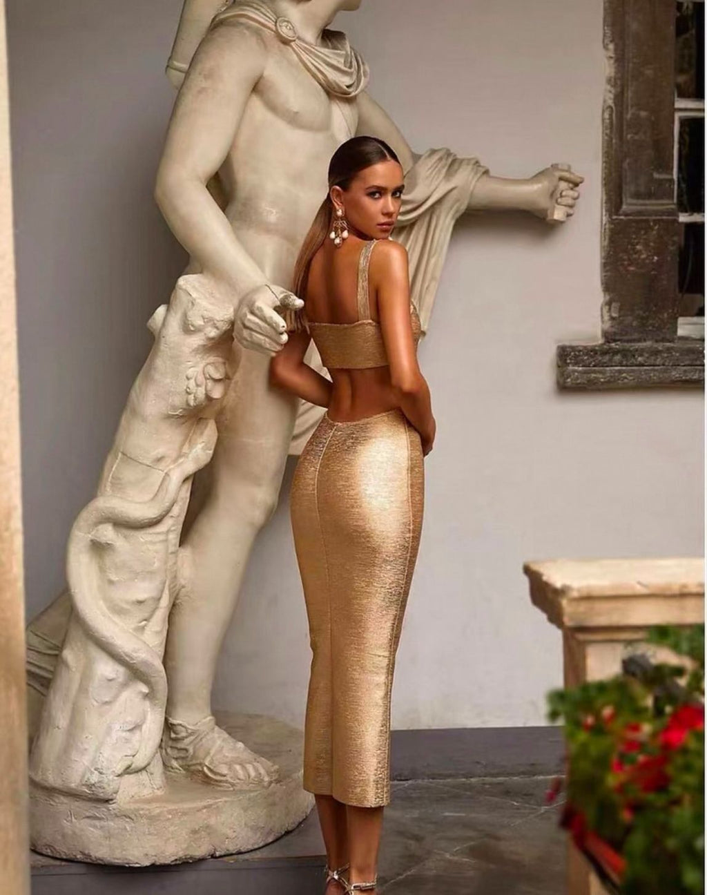 This image features a woman standing in a museum setting, beside a classical statue, which adds an elegant and timeless touch to the display. She is wearing a striking gold maxi dress that boasts a shimmering metallic texture, accentuating a sophisticated and glamorous look.