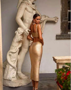 This image features a woman standing in a museum setting, beside a classical statue, which adds an elegant and timeless touch to the display. She is wearing a striking gold maxi dress that boasts a shimmering metallic texture, accentuating a sophisticated and glamorous look.