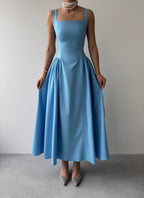 A woman wearing a blue sleeveless, square neckline, full-length dress with a pleated skirt.