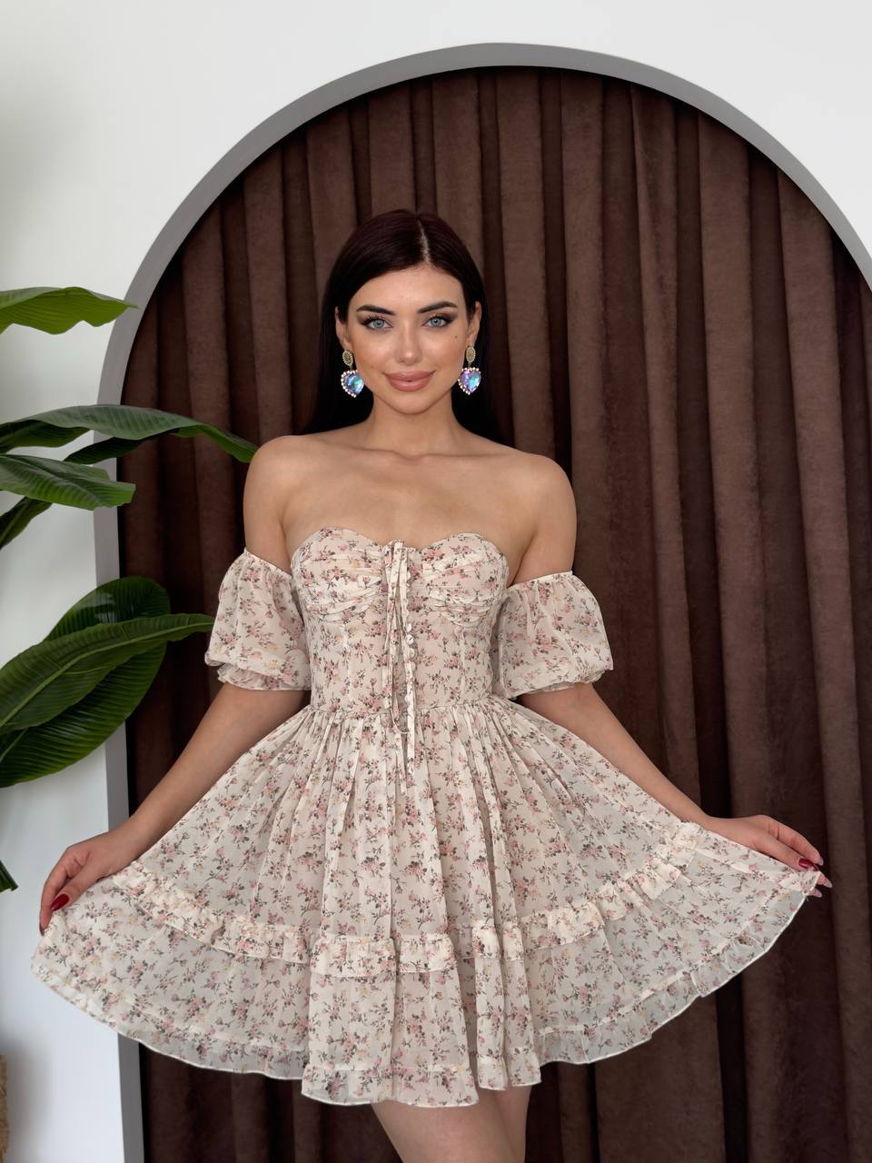 Woman in a floral dress standing in front of a curtain with plants.