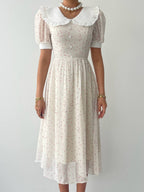 A close-up of a woman in a white dress with short puffed sleeves, featuring a round neckline with a ruffled collar and small red dot pattern. 