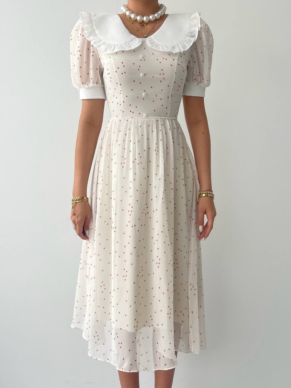 A close-up of a woman in a white dress with short puffed sleeves, featuring a round neckline with a ruffled collar and small red dot pattern. 