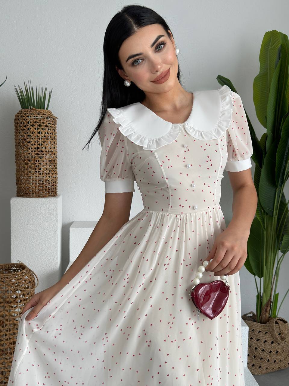 Woman in a white dress with a white collar, sitting indoors with plants around.