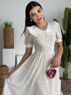 Woman in a white dress with a white collar, sitting indoors with plants around.