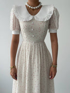 A close-up of a woman in a white dress with short puffed sleeves, featuring a round neckline with a ruffled collar and small red dot pattern. 