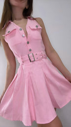A woman wearing a pink sleeveless dress with a belt and buttons on the front, and a pleated skirt.