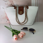 A white handbag with a brown leather trim and a chain shoulder strap, displayed alongside a pair of sunglasses and pink tulips, against a background with a pink floral pattern.