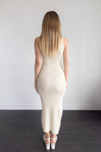 Woman wearing a beige dress standing against a plain white wall.